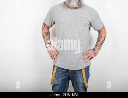 Nahaufnahme eines bärtig tätowierten Mannes in Jeans mit Hosenträgern und grauem Baumwoll-T-Shirt auf weißem Hintergrund. Stockfoto