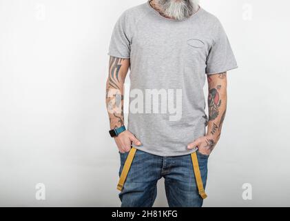 Bärtig tätowierte Mann posiert in Jeans mit Hosenträgern und grauem Kurzarm-T-Shirt aus Baumwolle, auf weißem Hintergrund. Stockfoto