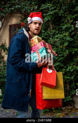 Weihnachtseinkäufe. Der junge Mann mit Weihnachtsmann-Hut hat einen überraschten Ausdruck und bringt seine neu gekauften Geschenke mit. Stockfoto