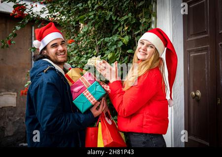 Weihnachtseinkäufe. Glückliches junges Paar beschäftigt mit Weihnachtsgeschenken außerhalb des Hauses. Er bringt viele Geschenkverpackungen und Umschläge, sie nimmt einen kleinen Stockfoto