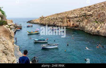 Menschen schwimmen im Meer an der Bucht Wied iż-Żurrieq zwischen Klippen, zwischen festgetäuten Booten und Luzzus - traditionelle bunte Holzfischerboote. Stockfoto