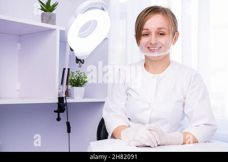 Nahaufnahme einer jungen, schönen professionellen Kosmetikerin, die lächelnd an einem Schreibtisch sitzt und im Schönheitssalon in Uniform und Medizin arbeitet Stockfoto