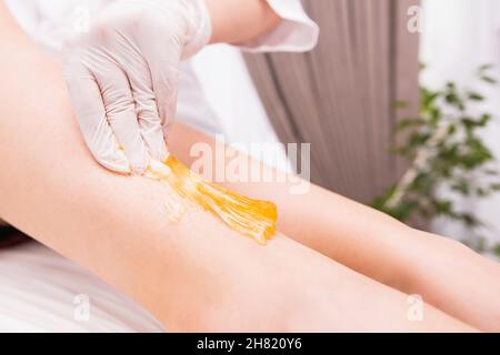 Frau, die in einem schönen Salon ein Haarentfernungsverfahren mit Zuckerpaste am Bein von einem Kosmetikerin mit weißen Schutzhandschuhen durchgeführt hat. Stockfoto