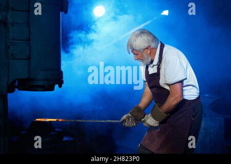 Seitenansicht des kaukasischen grauhaarigen Schmieds in Schutzuniform, der bei der Schmiede mit geschmolzenem Metall arbeitet. Handwerker, der Stahl unter der Pressenmaschine biegt. Stockfoto