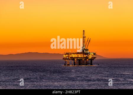 Offshore-Ölplattform vor der Küste Kaliforniens gegen einen launischen, orangen Himmel, wenn die Sonne hinter der Bohrinsel untergeht. Stockfoto