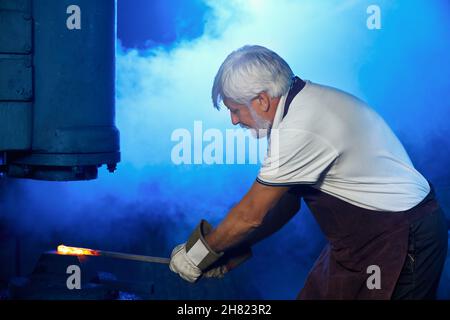 Kaukasischer Mann mit Schutzhandschuhen und Schürze, die beheizten Stahl mit einer Zepel unter der Pressenmaschine hält. Professioneller Schmied, der bei FORGE Metall formt. Stockfoto