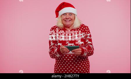 Großmutter in Weihnachten Pullover suchen Smartphone-Display aufrichtig jubeln Erfolg Glück zu gewinnen Stockfoto