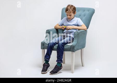 Kiew, Ukraine, Oktober 9 2021: Kleiner Junge hält Rubiks Würfel und spielt damit. Kind sitzt in einem gemütlichen Sessel und löst mentale Rätsel. Stockfoto