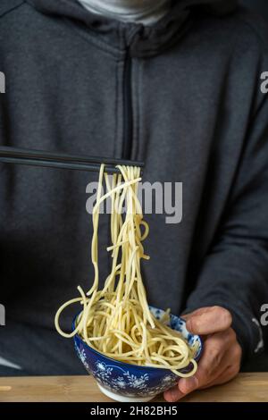 Spaghetti mit Essstäbchen halten und mit den Händen eines Jungen aus einer blauen Schüssel ziehen Stockfoto