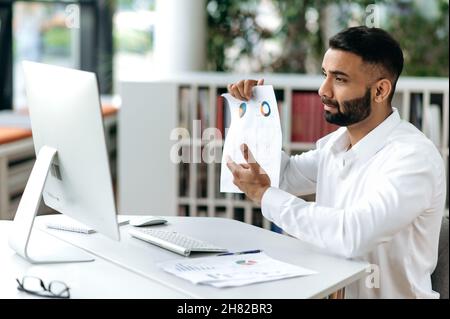Intelligenter erfolgreicher seriöser indischer Geschäftsmann oder CEO mit Bart im Büro sitzend, Computer verwendend, über Videokommunikation mit Geschäftspartnern oder Angestellten sprechend, Dokumente und Diagramme vorzeigend Stockfoto