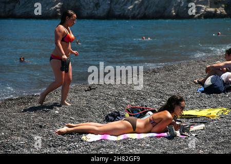 SANTORINI, GRIECHENLAND - 30. JUNI: Sonnenbaden und Schwimmen am schwarzen Strand von Kamari am 30. Juni 2012 in Santorini, Griechenland. Der Strand ist mit feinem schwarzen Sand bedeckt und fällt scharf ins Wasser ab. Stockfoto