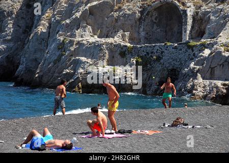 SANTORINI, GRIECHENLAND - 30. JUNI: Sonnenbaden und Schwimmen am schwarzen Strand von Kamari am 30. Juni 2012 in Santorini, Griechenland. Der Strand ist mit feinem schwarzen Sand bedeckt und fällt scharf ins Wasser ab. Stockfoto