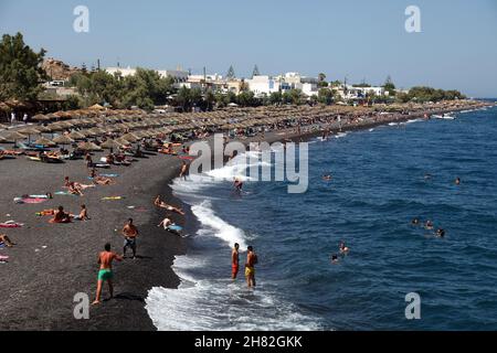 SANTORINI, GRIECHENLAND - 30. JUNI: Sonnenbaden und Schwimmen am schwarzen Strand von Kamari am 30. Juni 2012 in Santorini, Griechenland. Der Strand ist mit feinem schwarzen Sand bedeckt und fällt scharf ins Wasser ab. Stockfoto