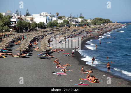 SANTORINI, GRIECHENLAND - 30. JUNI: Sonnenbaden und Schwimmen am schwarzen Strand von Kamari am 30. Juni 2012 in Santorini, Griechenland. Der Strand ist mit feinem schwarzen Sand bedeckt und fällt scharf ins Wasser ab. Stockfoto