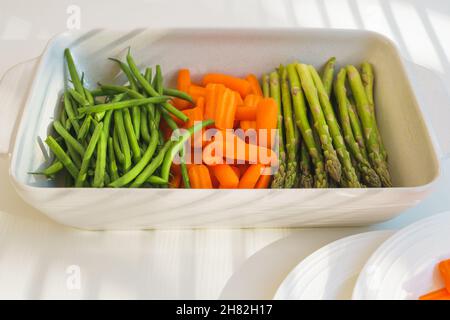 Rohe grüne Bio-Bohnen, Babykarotten, grüner Spargel in der Backform, fertig zum Kochen. Nahaufnahme, weißer Küchentisch-Hintergrund mit hellem li Stockfoto