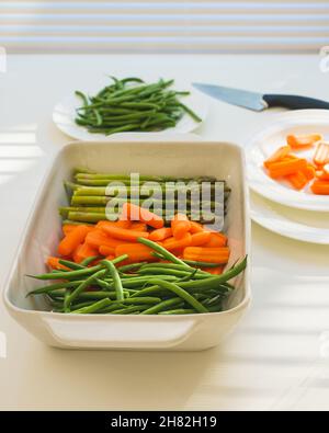 Rohe grüne Bio-Bohnen, Babykarotten, grüner Spargel in der Backform, fertig zum Kochen. Nahaufnahme, weißer Küchentisch-Hintergrund mit hellem li Stockfoto