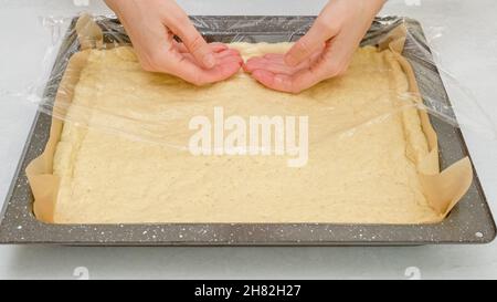 Die Hände der Frau mit Plastikfolie entrollen den Pizzateig auf die vorbereitete Backform. Hausgemachte Pizza Schritt für Schritt Rezept Stockfoto