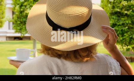 Frau in einem Strohhut hält eine Tasse heißes Getränk und trinkt Kaffee, sitzt am Morgen auf einer Sommerterrasse an einem sonnigen Tag. Das Konzept des Guten Stockfoto
