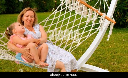 Das fröhliche Paar Großmutter und Enkelin 3 Jahre alt entspannen sich und sonnen sich in einer Hängematte auf einer grünen Wiese in ihrem Garten an einem sonnigen Sommer Stockfoto