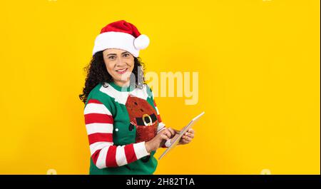 Porträt einer erwachsenen lateinerin mit Tablet mit Kopierfläche in einem weihnachtskonzept auf gelbem Hintergrund Stockfoto