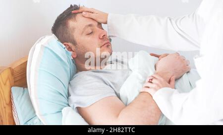 Arzt überprüft die Fiebertemperatur eines älteren Mannes, der auf dem Bett liegt. Mann, der sich zu Hause ausruht und Symptome der Grippe fühlt, während Frau Fieber durch Berühren von f überprüft Stockfoto