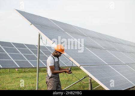 Professioneller afrikanischer Ingenieur in Helm und Uniform mit Multimeter zur Messung der Stromstärke in Solarmodulen. Konzept von Menschen, Wartung und alternativer Energie. Stockfoto