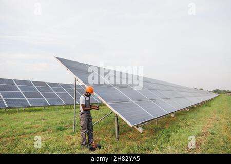 Professioneller afrikanischer Ingenieur in Helm und Uniform mit Multimeter zur Messung der Stromstärke in Solarmodulen. Konzept von Menschen, Wartung und alternativer Energie. Stockfoto