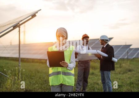 Die muslimische Frau hijab schrieb auf die Zwischenablage, während ihre männlichen Kollegen hinter Helmen standen und Blaupausen untersuchten. Multirassische Menschen, die die Produktion von Sonnenenergie entwickeln. Stockfoto