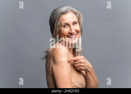 Reife grauhaarige Frau schützt sich mit einem Impfstoff gegen Covid und andere Viruserkrankungen, glückliche ältere Frau zeigt mit dem Finger auf einen medizinischen Fleck auf ihrer Armschulter. Gesundheitswesen und Medizin Stockfoto
