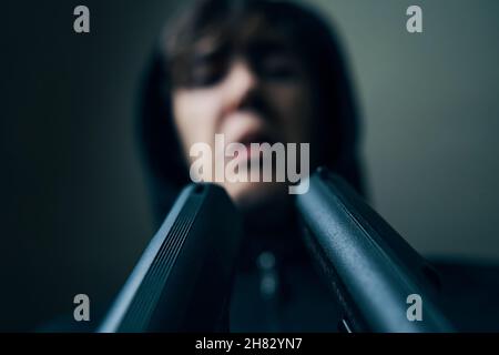 Zwei Gewehre zeigten auf das Gesicht des Menschen. Nahaufnahme von Schusswaffen. Verhör eines Mannes in einer Kapuze mit Pistolen. Drohungen und Einschüchterung. Stockfoto