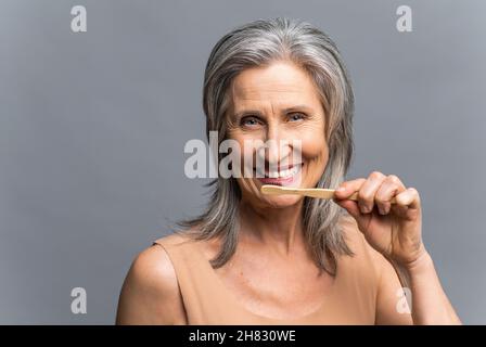 Kopfaufnahme einer lächelnden älteren grauhaarigen Frau mit Bambuszahnbürste, die ihre weißen Zähne auf grauem Hintergrund putzt, tägliche Pflegeroutine. Zahnpflege und Mundhygiene Stockfoto