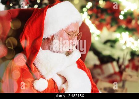 Porträt des Weihnachtsmanns mit einem echten grauen Bart, der auf einem Stuhl vor dem Hintergrund eines Weihnachtsbaums und Weihnachtslichtern sitzt. Stockfoto