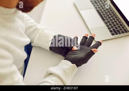 Mann mit rheumatoider Arthritis trägt Kompressionshandschuhe zur Schmerzlinderung, während er am Laptop arbeitet Stockfoto