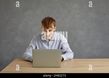 Millennial Mann sitzt auf grauem Hintergrund auf einem Laptop online an der Schreibtischarbeit Stockfoto