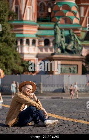 Junger männlicher Reisender in braunem Hut sitzt auf dem Hintergrund des Roten Platzes Russland. Stockfoto
