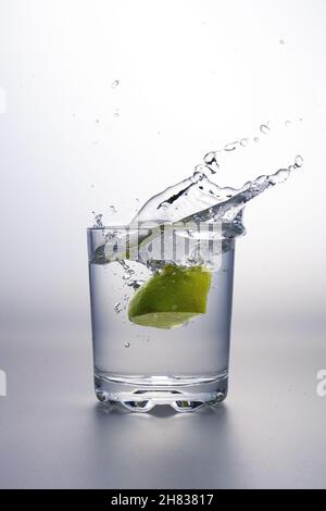 Kalkbrocken, die auf einer grauen Oberfläche und weißem Hintergrund in ein Glas mit klarer Flüssigkeit oder Wasser spritzt Stockfoto
