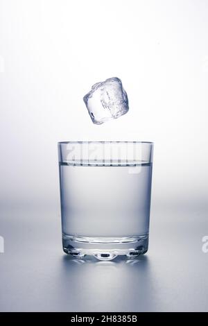 Glas klares Wasser mit einem Eiswürfel, der darüber fällt. Auf grauer Oberfläche und weißem Hintergrund. Stockfoto