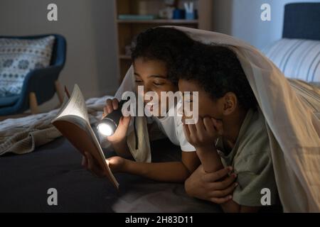 Positive afroamerikanische Mädchen leuchtet Taschenlampe auf Buch, während es zu Bruder in der Dunkelheit zu lesen Stockfoto