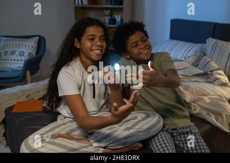 Fröhliche afroamerikanische Geschwister, die auf dem Bett sitzen und die Hände gegen das Licht der Taschenlampe gestikulieren, während sie den Schattenwurf der Tiere erzeugen Stockfoto