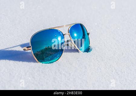 Sonnenbrillen mit blauen Linsen, in denen der Wald reflektiert wird, liegen auf dem Schnee Stockfoto