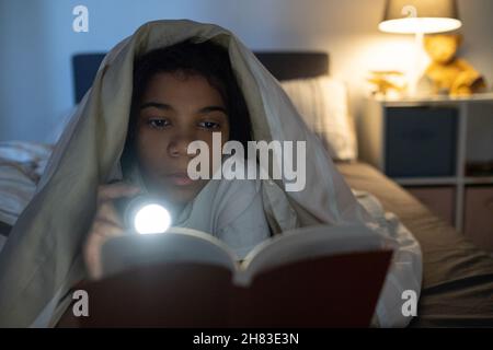 Seriöse afroamerikanische Mädchen unter Bettdecke auf dem Bett liegen und leuchtende Taschenlampe auf Buch in der Dunkelheit Stockfoto