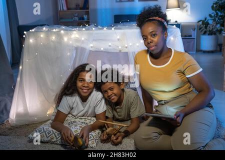 Portrait einer sorgfältigen afroamerikanischen Mutter mit Tablet, die am handgefertigten Zelt sitzt und Kindern Märchen liest Stockfoto