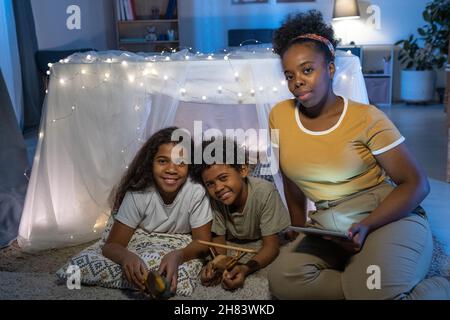 Portrait einer sorgfältigen afroamerikanischen Mutter mit Tablet, die am handgefertigten Zelt sitzt und Kindern Märchen liest Stockfoto