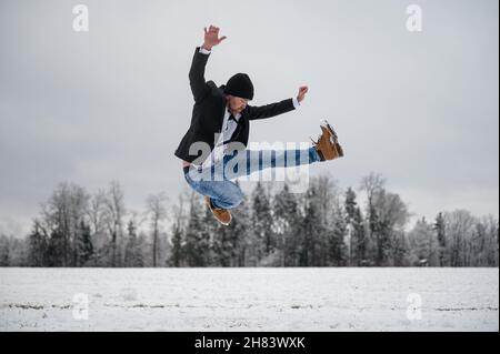 Fröhlicher, fröhlicher junger Mann in Jeans und Jacke, der vor Freude draußen in einer weiß verschneiten Natur springt. Stockfoto