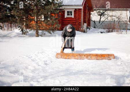 Russische Tradition der Teppichreinigung. Kaukasische ältere Frau reinigt Teppich mit Neuschnee mit Besen im Hinterhof am Wintertag Stockfoto
