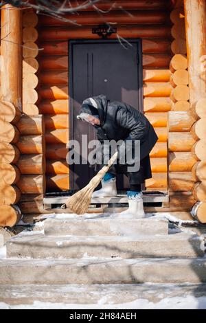 Kaukasische Frau reinigt Schnee. Ältere Frau in Daunenjacke und Filzstiefeln fegt Schnee von der Veranda mit Besen auf dem Hintergrund des Holzhauses Stockfoto