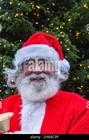 London, Großbritannien. 27. November 2021. Ein Mann, der als traditioneller Weihnachtsmann (Weihnachtsmann) gekleidet ist, steht mit einer Glocke vor einem Weihnachtsbaum im Stadtzentrum von Wimbledon, während die Menschen mit weniger als einem Monat bis Weihnachten in den festlichen Geist eintauchen. Kredit: amer ghazzal/Alamy Live Nachrichten Stockfoto