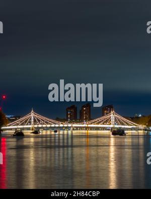 Die Albert Bridge wurde nachts mit Wolkenkratzern im Hintergrund und der themse im Vordergrund beleuchtet. Aufnahme von der Chelsea Bridge. Stockfoto