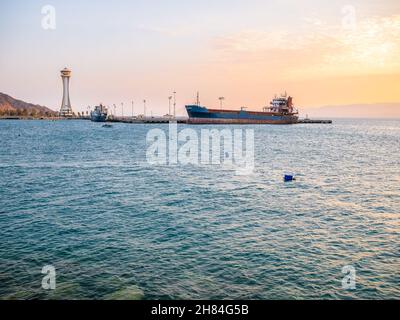 Panoramablick bei Sonnenuntergang mit einem angedockten alten verrosteten Schiff und dem Aussichtsturm des Leuchtturms im Hafen von Aqaba, Jordanien. Stockfoto