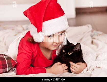 Kleines Mädchen in lustigen Weihnachtsmütze und Weihnachts-Pyjamas liegt auf einem Karo mit Lieblings-Haustier schwarze Katze. Stockfoto